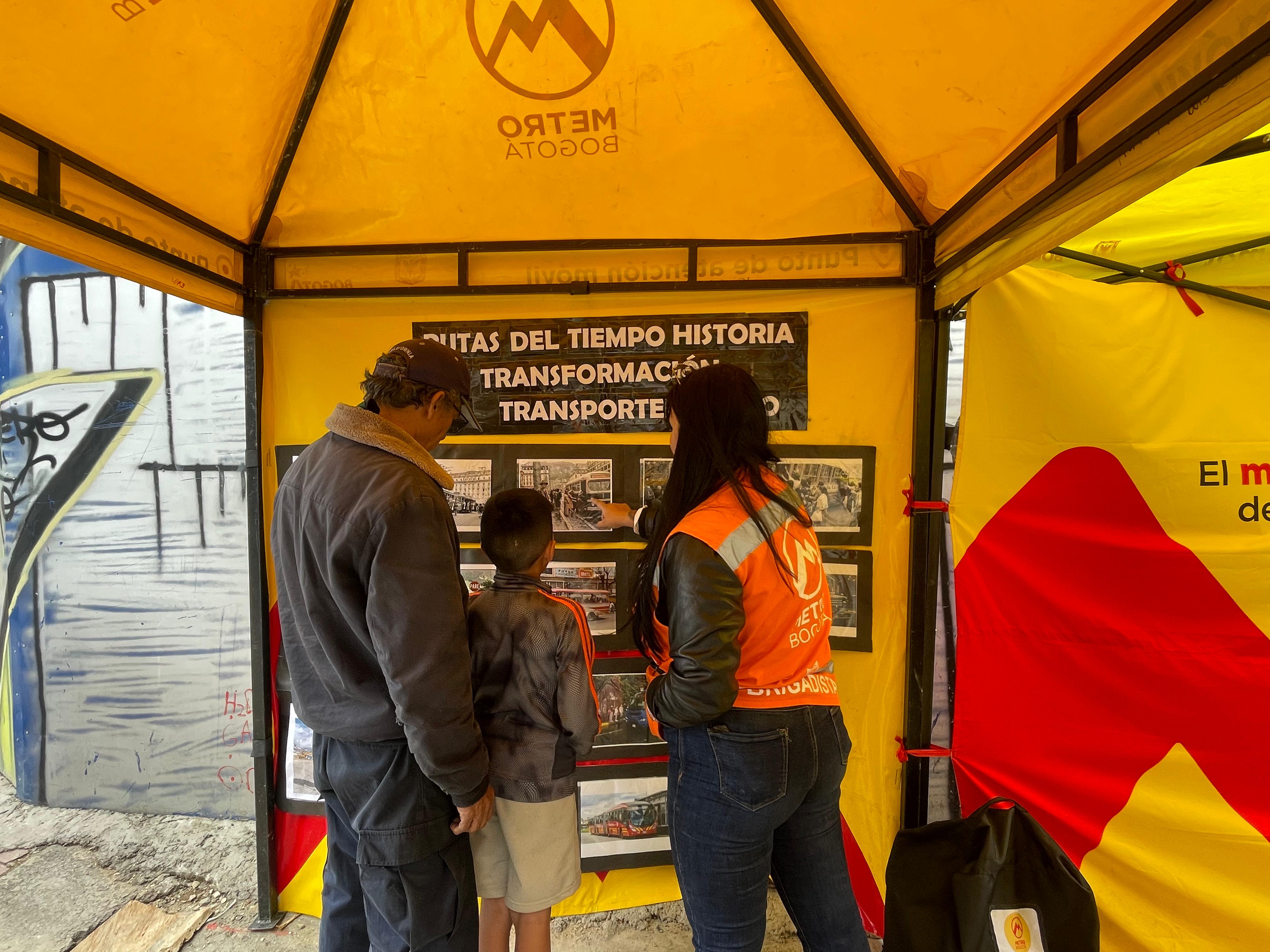 En una carpa amarilla de atención al ciudadano, una mujer con chaleco del Metro le muestra a un niño y su padre las fotos de una activación pedagógica que se hizo para las obras del Metro