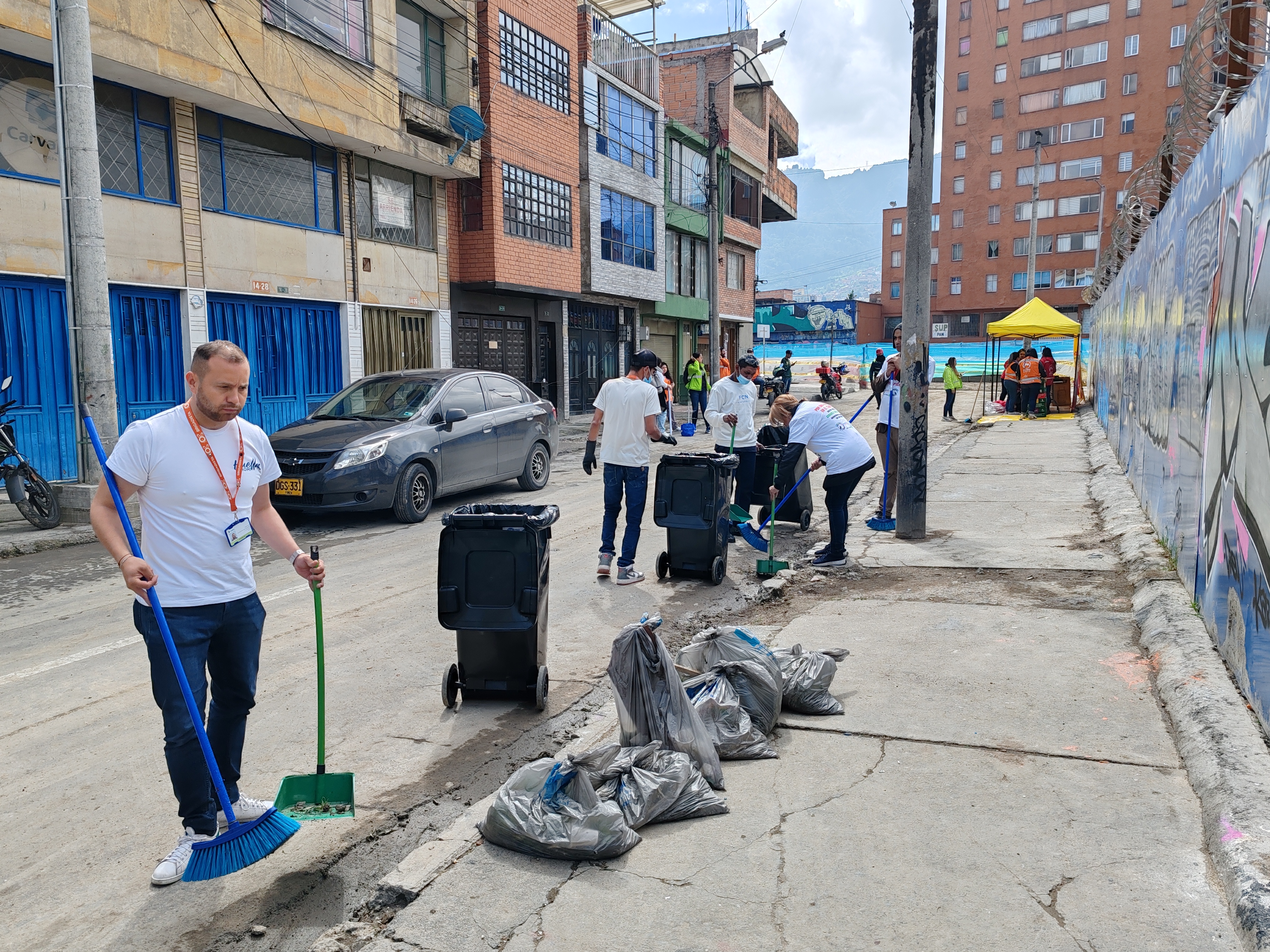 Varias personas en una calle del barrio, todas con camiseta blanca barriendo las calles, limpiando las zonas comunes de la zona, tienen recogedores grandes de basura y el espacio se ve limpio